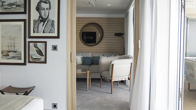Owner's Suite aboard Le Bellot luxury expedition ship, showing king bed, separate living room, bright white decor & photos & drawings of explorers on the walls.