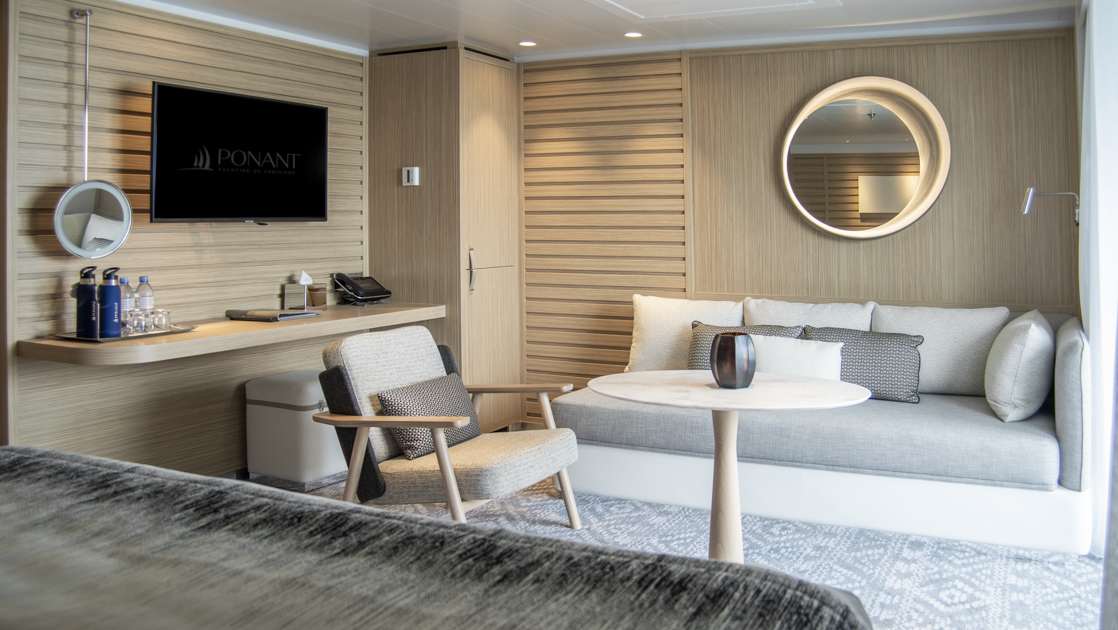 Privilege Suite aboard French luxury ship Le Bellot, with king bed, couch, table, chair & private balcony, plus natural-looking decor.