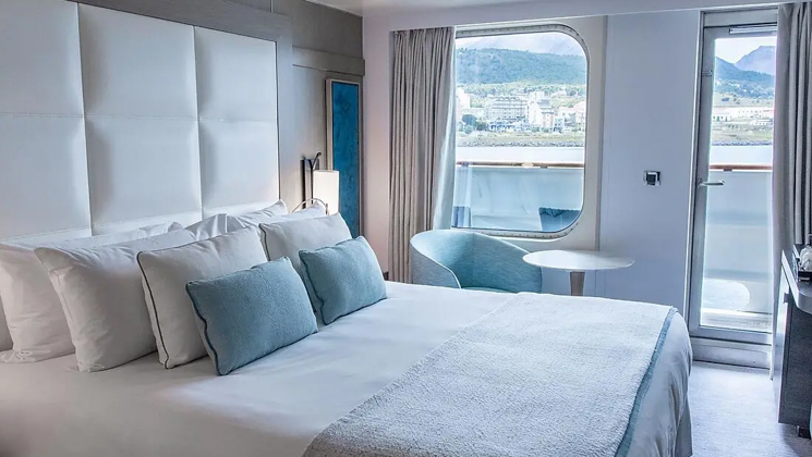 Deluxe stateroom aboard Le Boreal expedition ship, showing king bed, table, chair & white-&-teal appointments.