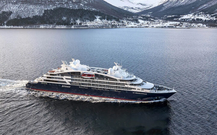 Le Bougainville cruise ship with blue hull, curved white upper decks & red accents sails past snowy mountains.