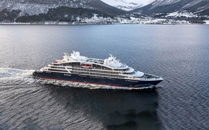 Le Bougainville cruise ship with blue hull, curved white upper decks & red accents sails past snowy mountains.