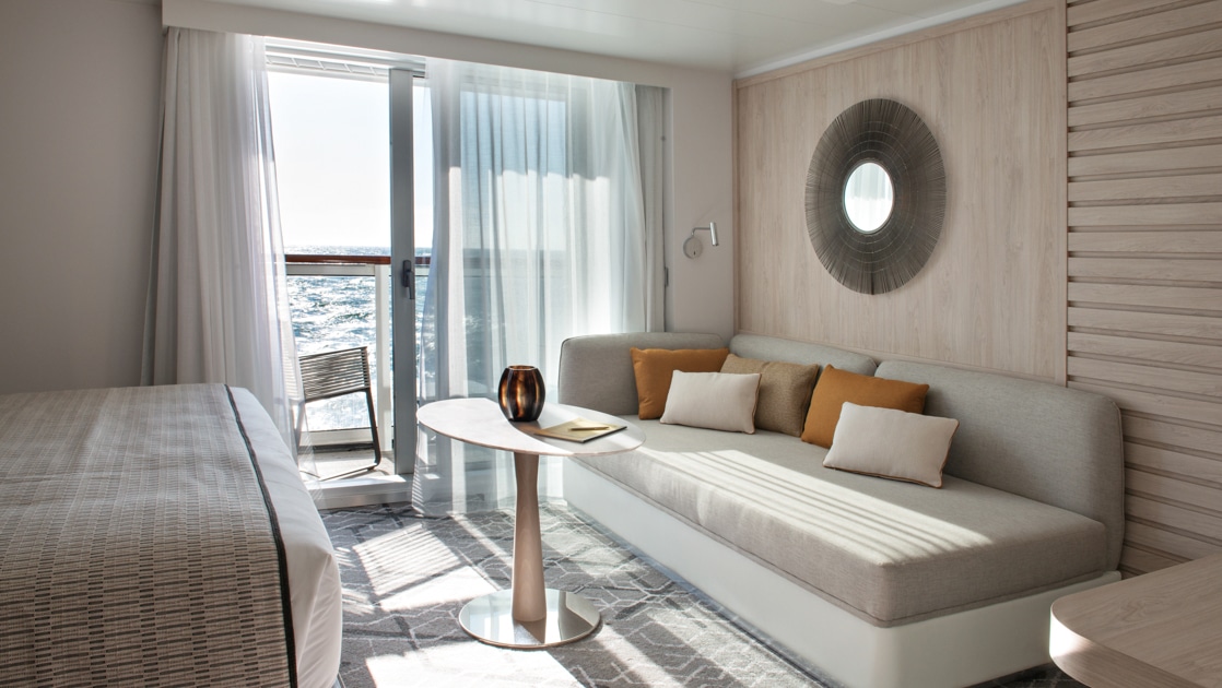 View from bed to couch beside window doors to balcony in the beige-accented Deluxe Suite on Le Bougainville cruise ship.