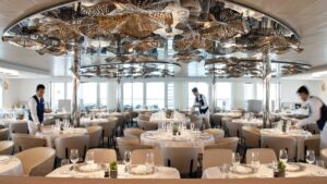 Dining room with elegant white tablecloths & floral gold light fixtures, being set for dinner by servers aboard Le Bougainville.