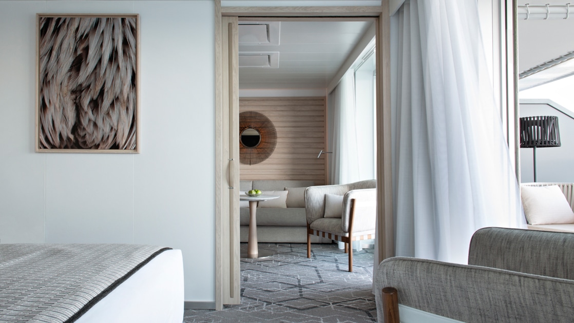 View from bedroom to window-lined living room with couch in the beige-accented Grand Deluxe Suite on Le Bougainville ship.
