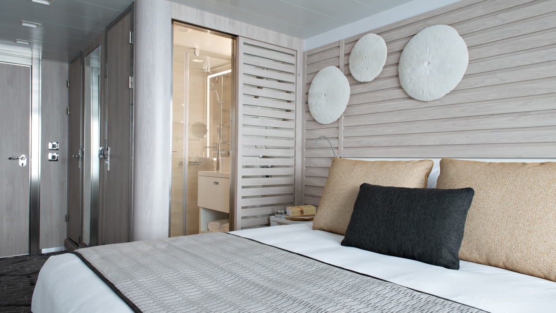 View towards bathroom, entryway & door from double bed in beige-accented Prestige Stateroom on Le Bougainville ship.