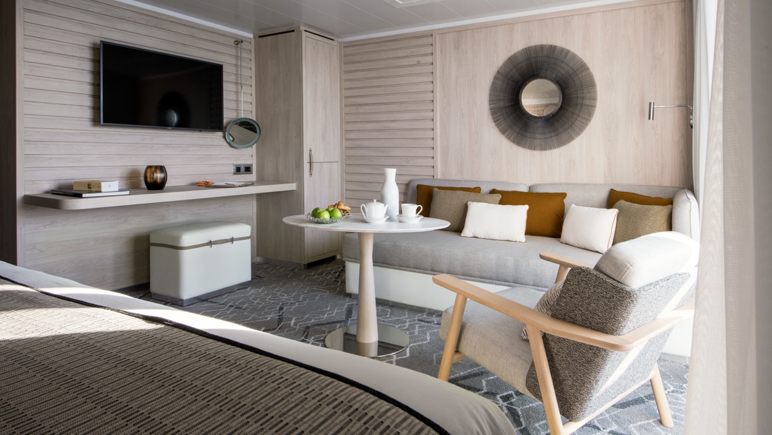 View from bed beside living room with couch, desk & TV in the beige-accented Owner's Suite on Le Bougainville cruise ship.