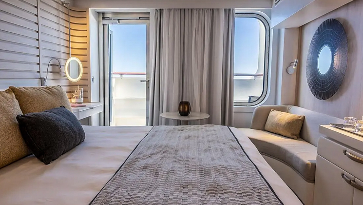 View from bed to dresser & couch beside window & door to balcony in the blue-accented Deluxe Stateroom on Le Bougainville ship.
