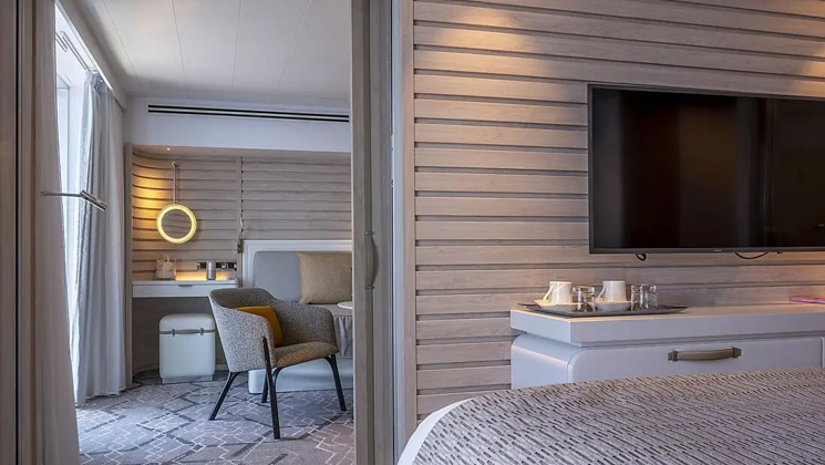 Double bed looks into adjoining room with couch & chair in beige-accented Prestige Suite on Le Bougainville ship.