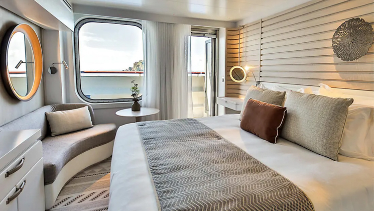 Deluxe Stateroom aboard Le Dumont D'Urville French ship, with king bed, white dresser, private balcony, chair & ethinc-yet-chic decor.