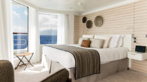 Grand Deluxe Suite aboard luxury French ship Le Dumont D'Urville, with ethnic chic decor, wooden headboard, king bed & wraparound windows.