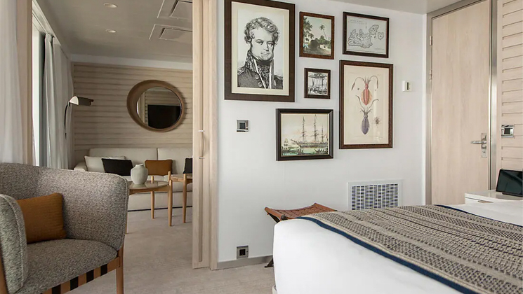 Owner's Suite aboard Le Dumont D'Urville luxury expedition ship, showing king bed, separate living room, bright white decor & photos & drawings of explorers on the walls.