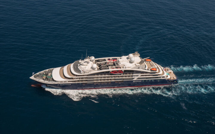 Aerial view of Le Jacques Cartier luxury expedition ship cruising in open waters, with dark blue & red hull & white upper decks.