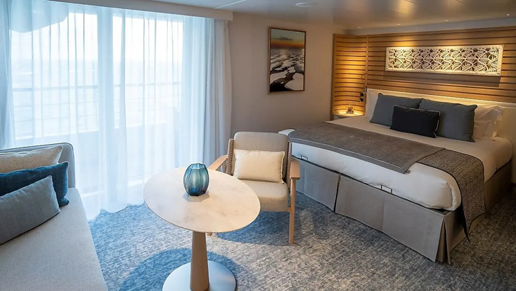 Privilege Suite aboard French luxury ship Le Jacques Cartier, with king bed, couch, table, chair & private balcony, plus natural-looking decor.