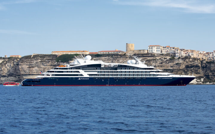 Le Laperouse small expedition ship with dark blue hull & white upper decks sits in calm blue sea beside cliffside town in sun.
