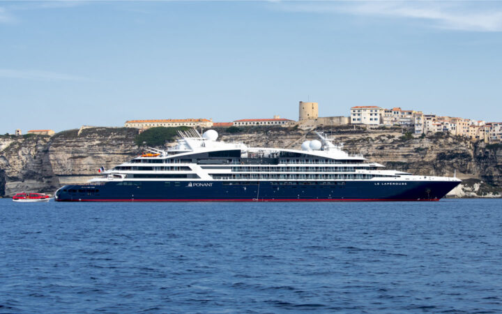 Le Laperouse small expedition ship with dark blue hull & white upper decks sits in calm blue sea beside cliffside town in sun.
