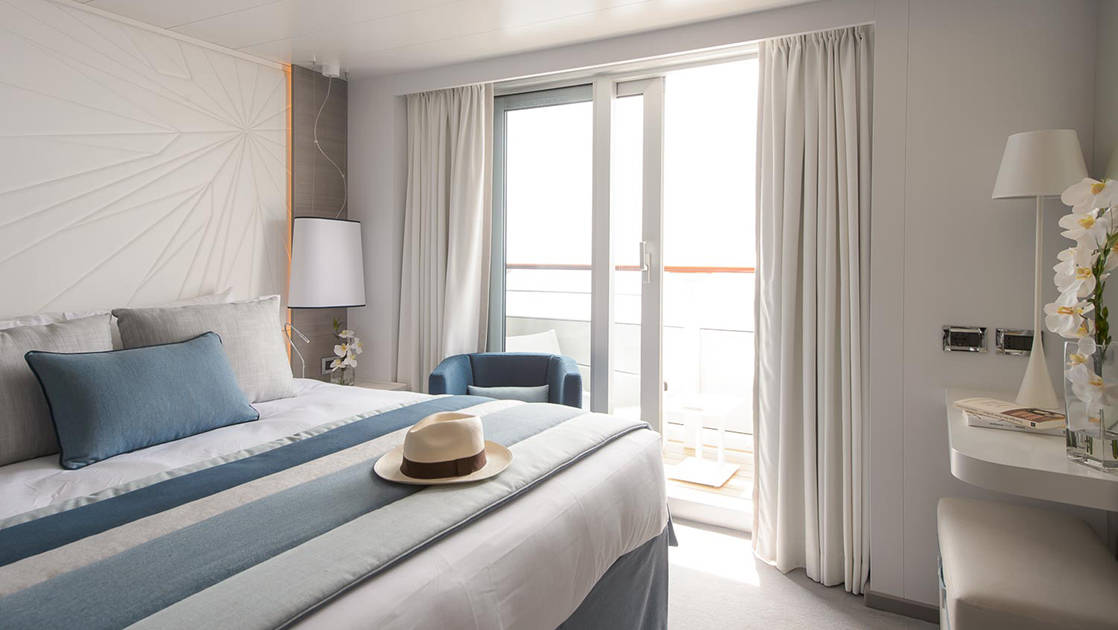 Grand Deluxe Suite on Le Lyrial French ship, with king bed, private balcony, desk & lamps, in white-&-blue decor.