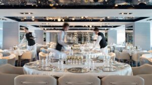 Main dining room aboard French luxury ship Le Lyrial, with white-&-ivory decor, silver place settings, white roses, wine glasses & servers setting empty tables.
