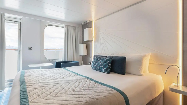 Deluxe Stateroom aboard Le Lyrial expedition ship, with blue-&-ivory decor, bed, bedside table & lamp & private balcony.