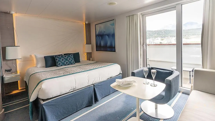 Deluxe Suite on Le Lyrial French ship, with king bed, private balcony, table, chair, couch & lamps, in white-&-blue decor.