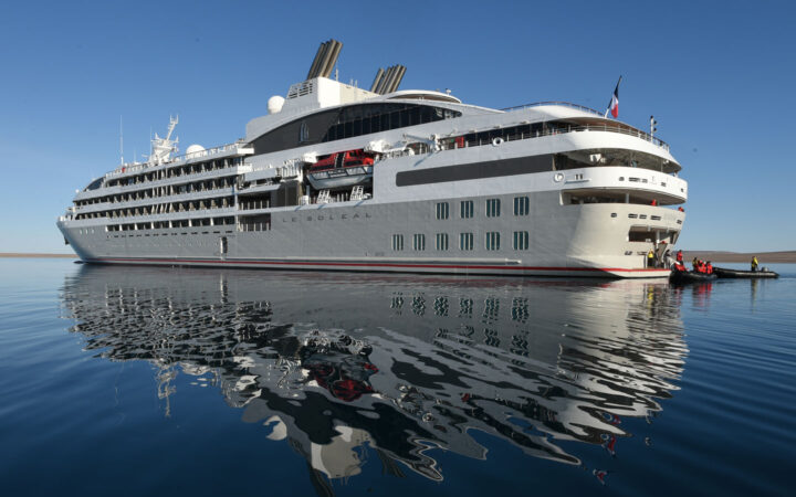 Luxury expedition ship Le Soleal foats on a blue body of water on a clear blue sky day, she is 7 stories high mainly white with a light grey hull her reflection is clear in the water.