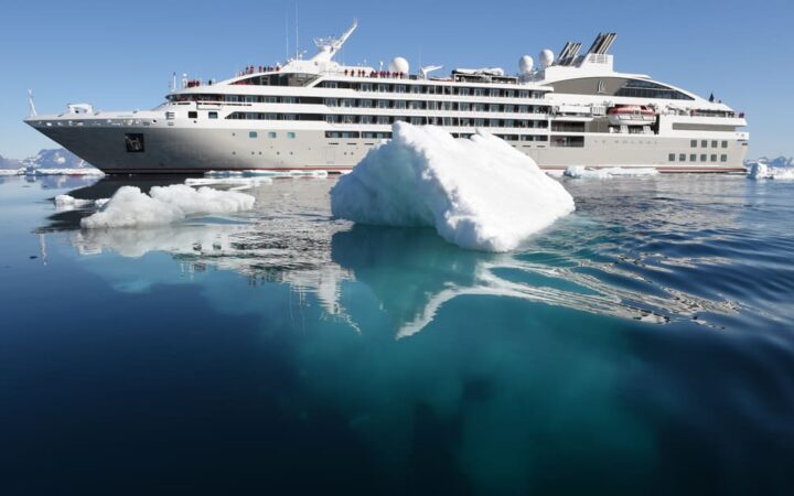 Le Soleal luxury small ship cruises Antarctica, in front of it is a small white iceberg, the clear teal water allows you to see the underside of the iceberg.