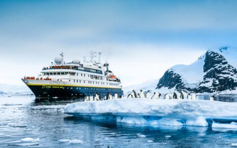 National Geographic Orion small ship with blue hull & white upper decks sits in calm sea by small iceberg with penguins.