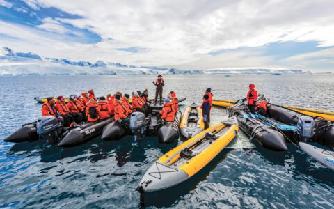 Travelers in orange jackets prepare to venture out in small skiffs and kayaks on a sunny and cloudy day