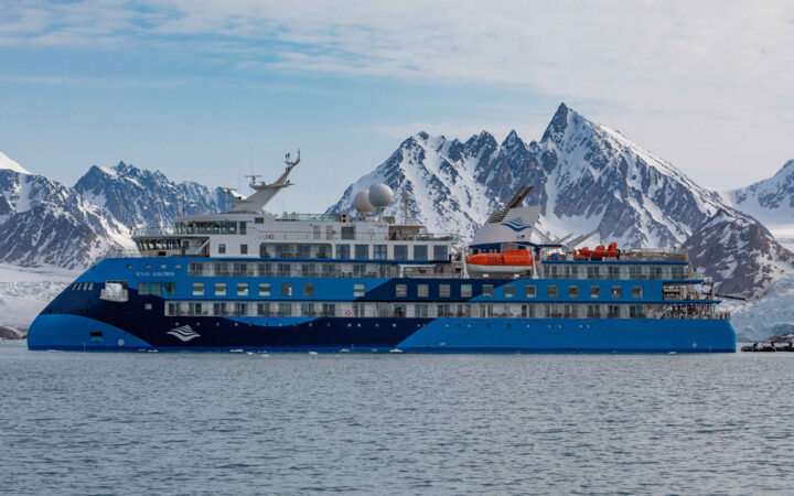 Exterior image of small ship cruise Antarctica with blue exterior in front of snowy mountains