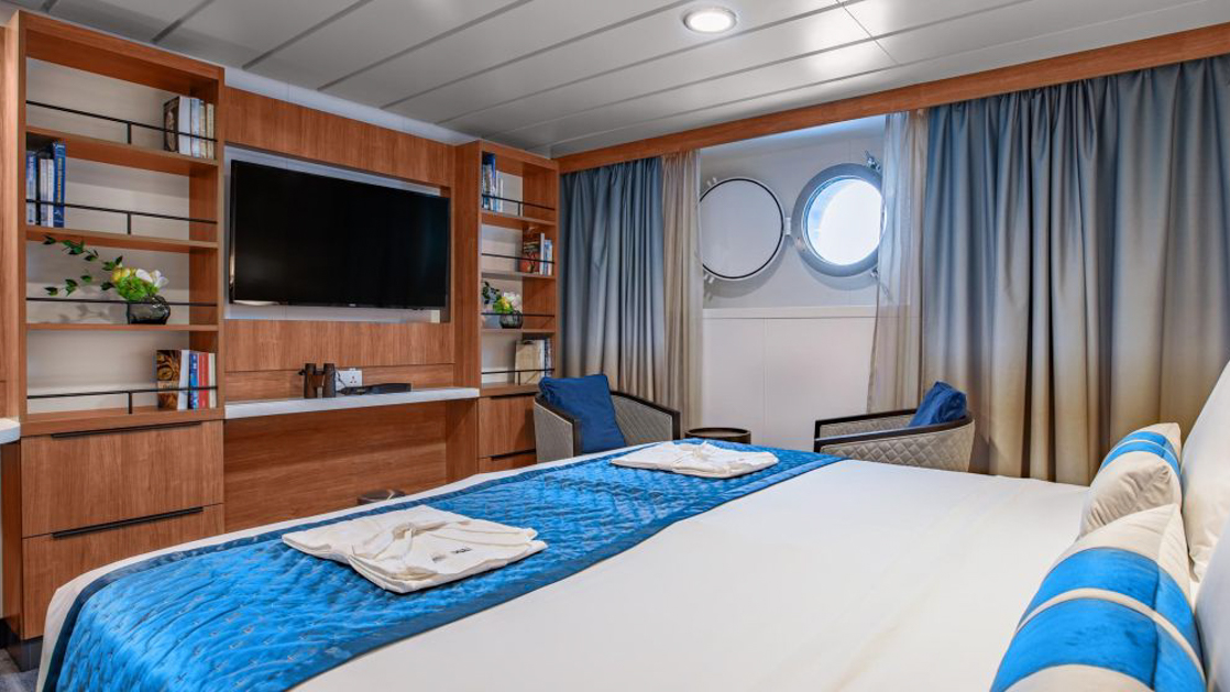 A modern cruise ship cabin with a television, and a window. The room has light wood paneling.