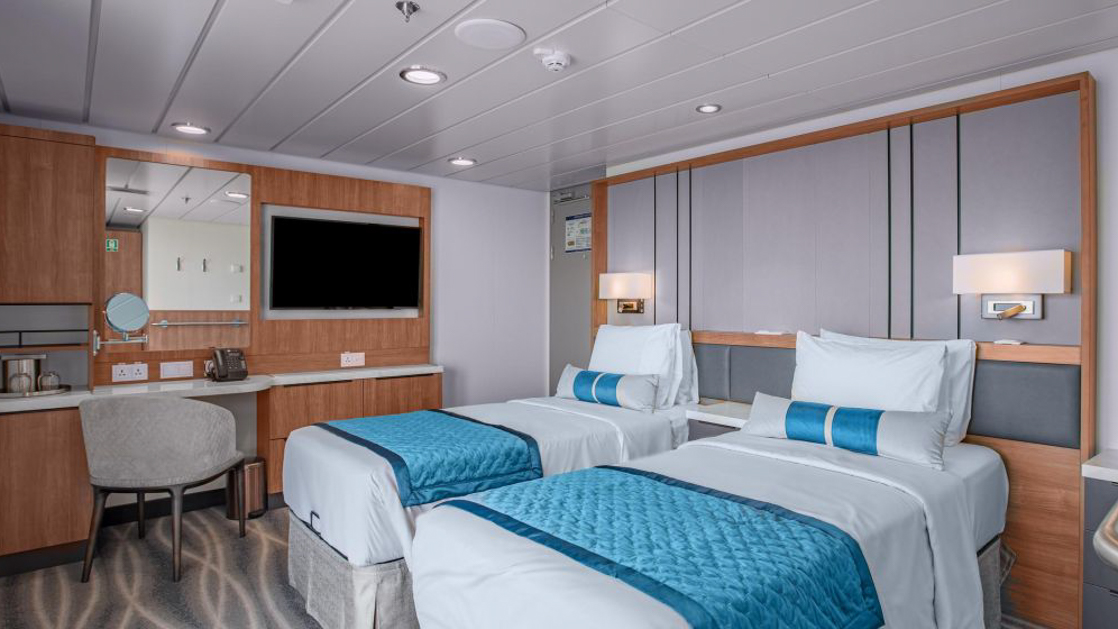 A modern cruise ship cabin with two single beds, a television, and a window. The room has light wood paneling.