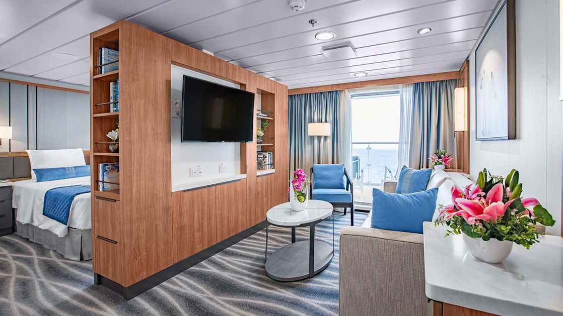 A modern cruise ship suite with a bedroom area, a living area with blue chairs, and a wooden partition with a TV.