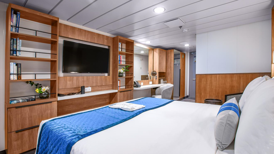 A modern cruise ship cabin with a double bed, a television, and a window. The room has light wood paneling.