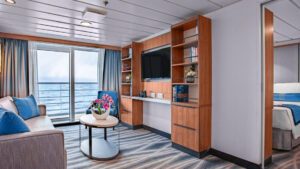 A modern cruise ship suite with a bedroom area, a living area with blue chairs, and a wooden partition with a TV.
