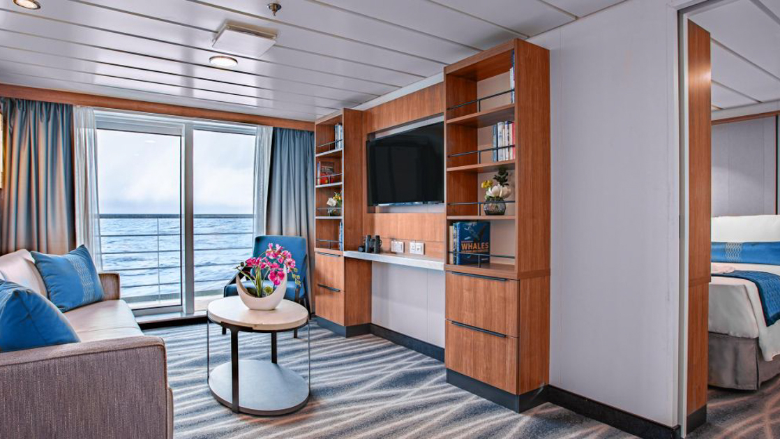 A modern cruise ship suite with a bedroom area, a living area with blue chairs, and a wooden partition with a TV.