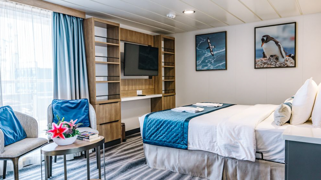 A modern cruise ship cabin with a double bed, a television, and a window. The room has light wood paneling.