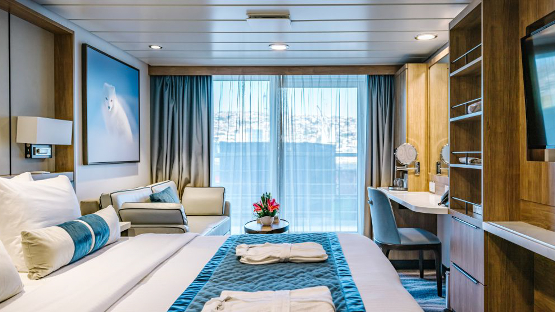 A modern cruise ship cabin with a double bed, a television, and a window. The room has light wood paneling.