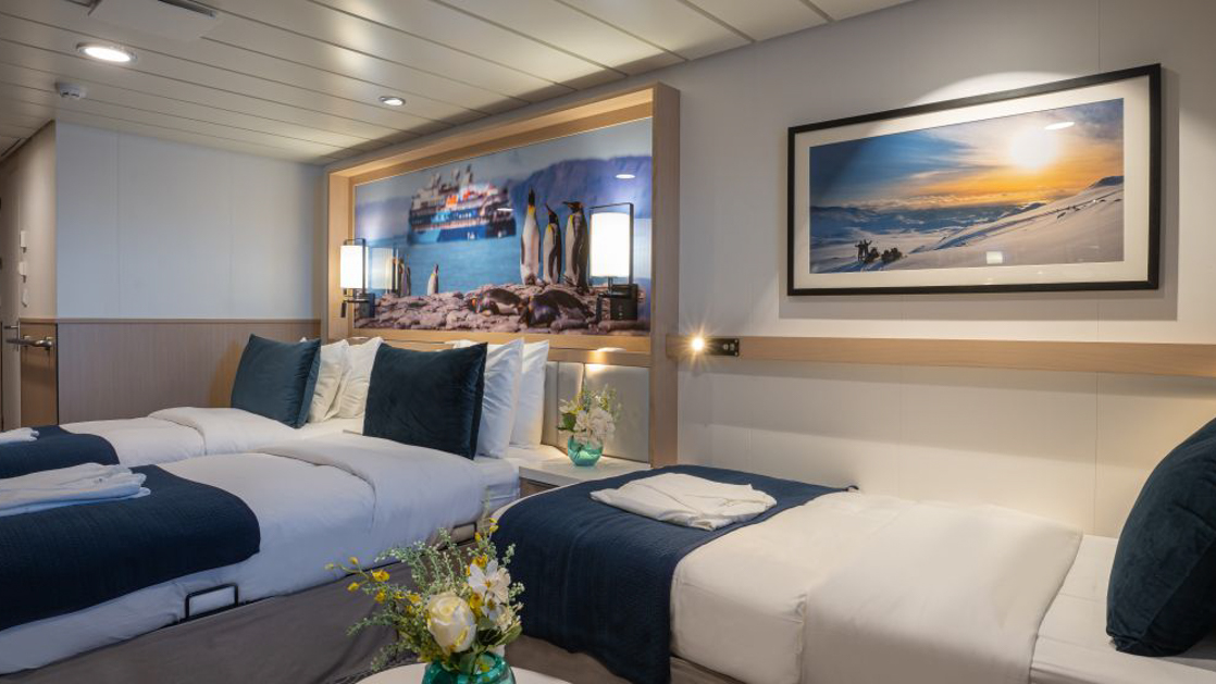 A modern cruise ship cabin with three single beds, a television, and a window. The room has light wood paneling.