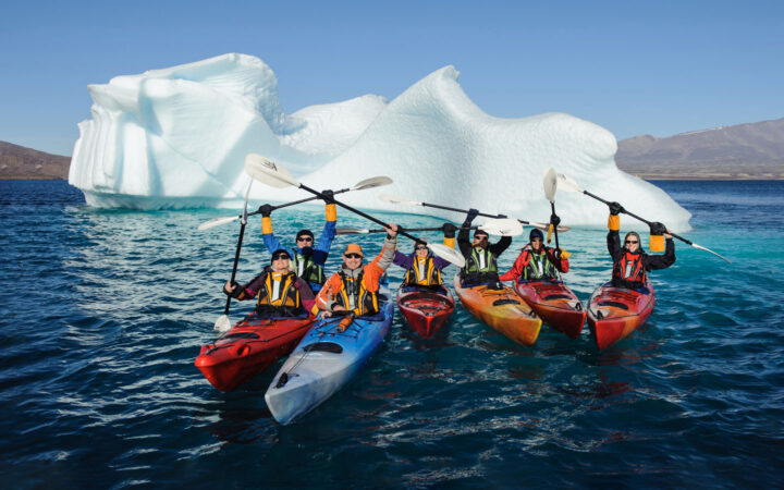 A group of kayakers in red orange and blue kayaks hold their paddles in the air as they float in front of a large iceberg in Antarctica