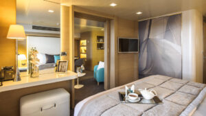 Owner's Suite aboard Le Soleal, a small vanity desk with a flat screen TV on the wall, a food tray sitn ontop of a grey comforter on the king bed