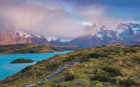 Aerial view of bright blue water beside snowcapped peaks & grassy hillside on a Patagonia to Antarctica cruise.