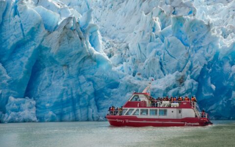 Red & white boat cruises beside a massive blue glacier in Torres del Paine National Park, Patagonia, Chile.