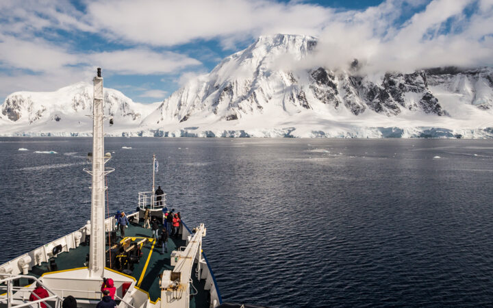 Polar Circle Cruise guests stand on bow of small green & white expedition ship look out at tall, snowy peaks in the sun.