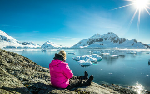 Woman in pink jacket sits on gray rock overlooking icy bay with glassy water & snowy peaks beyond on the Antarctic Peninsula.