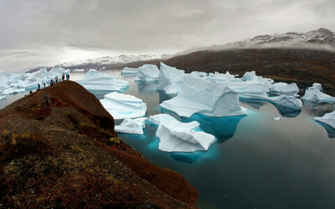 Hikers overlook large icebergs sitting in Arctic waters on a cloudy day during the Scoresby Sund Aurora Borealis voyage.