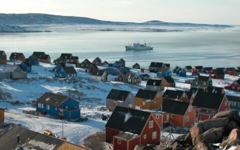 Aerial view of small seaside town in the Arctic with colorful homes & small ship sitting offshore, on a sunny day.