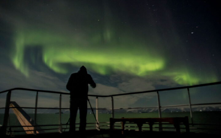 Person stands in deck of a ship at night, watching the green aurora borealis on the Scoresby Sund Aurora Borealis voyage.
