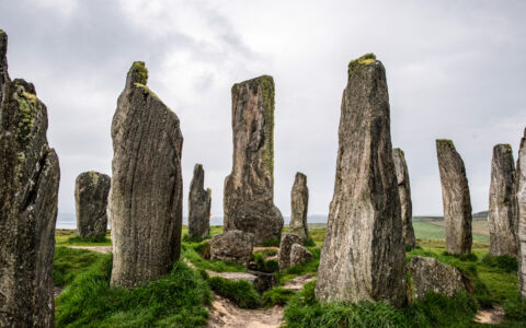 Ancient, tall Callanish stones stand in a ring under a cloudy sky with pathways & green grass beneath in Scotland.