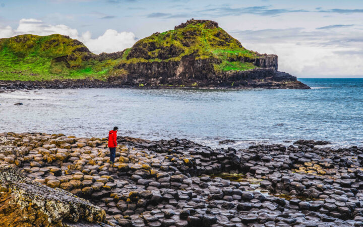 Scotland, Faroes & Iceland cruise guest in red jacket stands atop hexagonal stones near inlet & bright green hills.