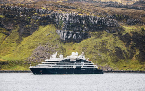Small blue & white expedition ship sits in calm fjord beside grassy green volcanic hills with dark stone cliffs in Iceland.