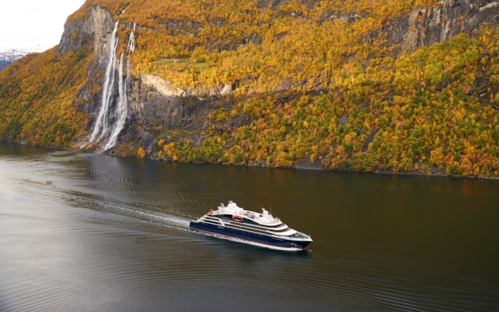 Small ship with blue hull & white upper decks cruises through calm Norwegian fjords with fall colors on hillside & waterfall.